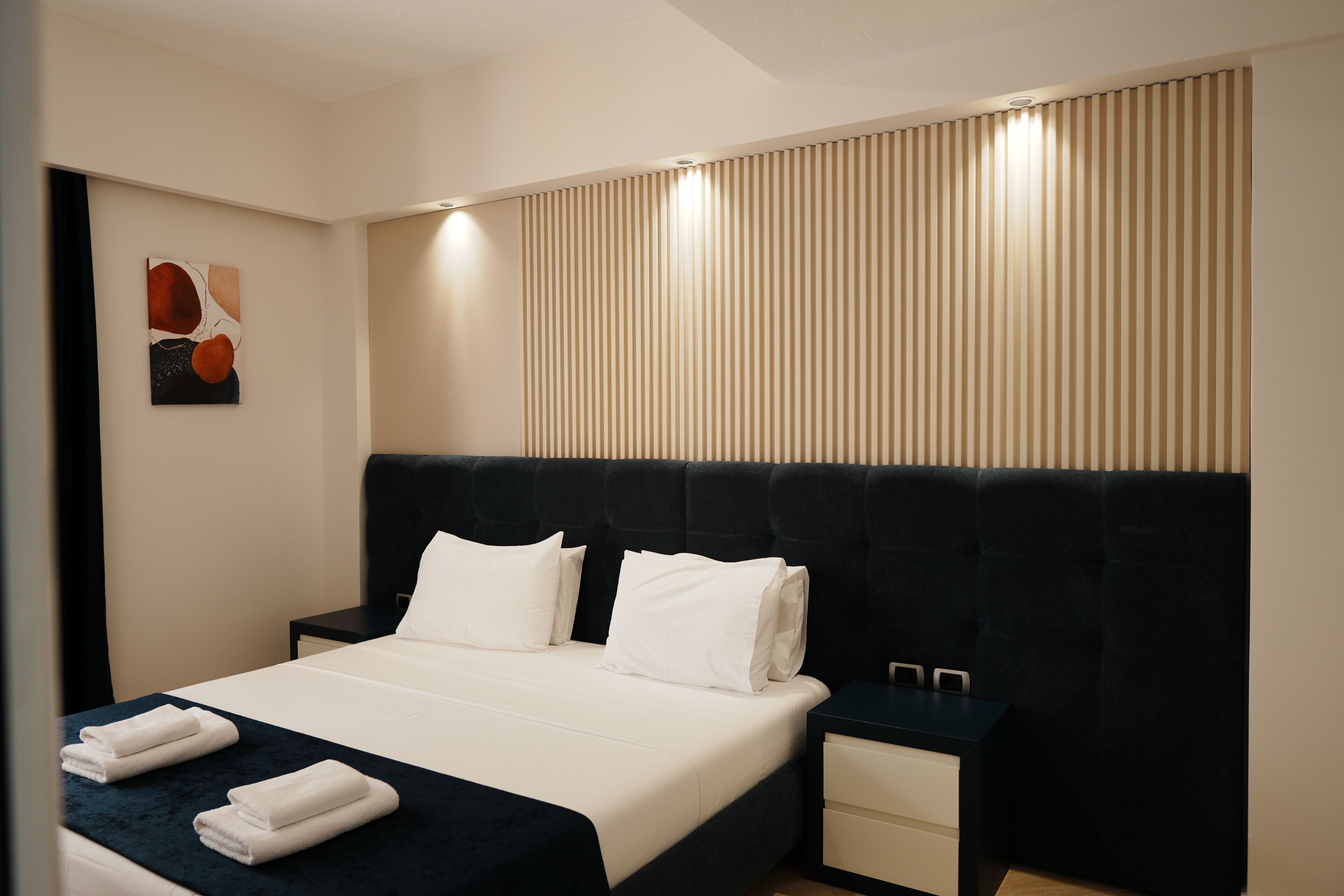 A modern hotel room with a large bed, bedside tables, a headboard, and a wall-mounted artwork.