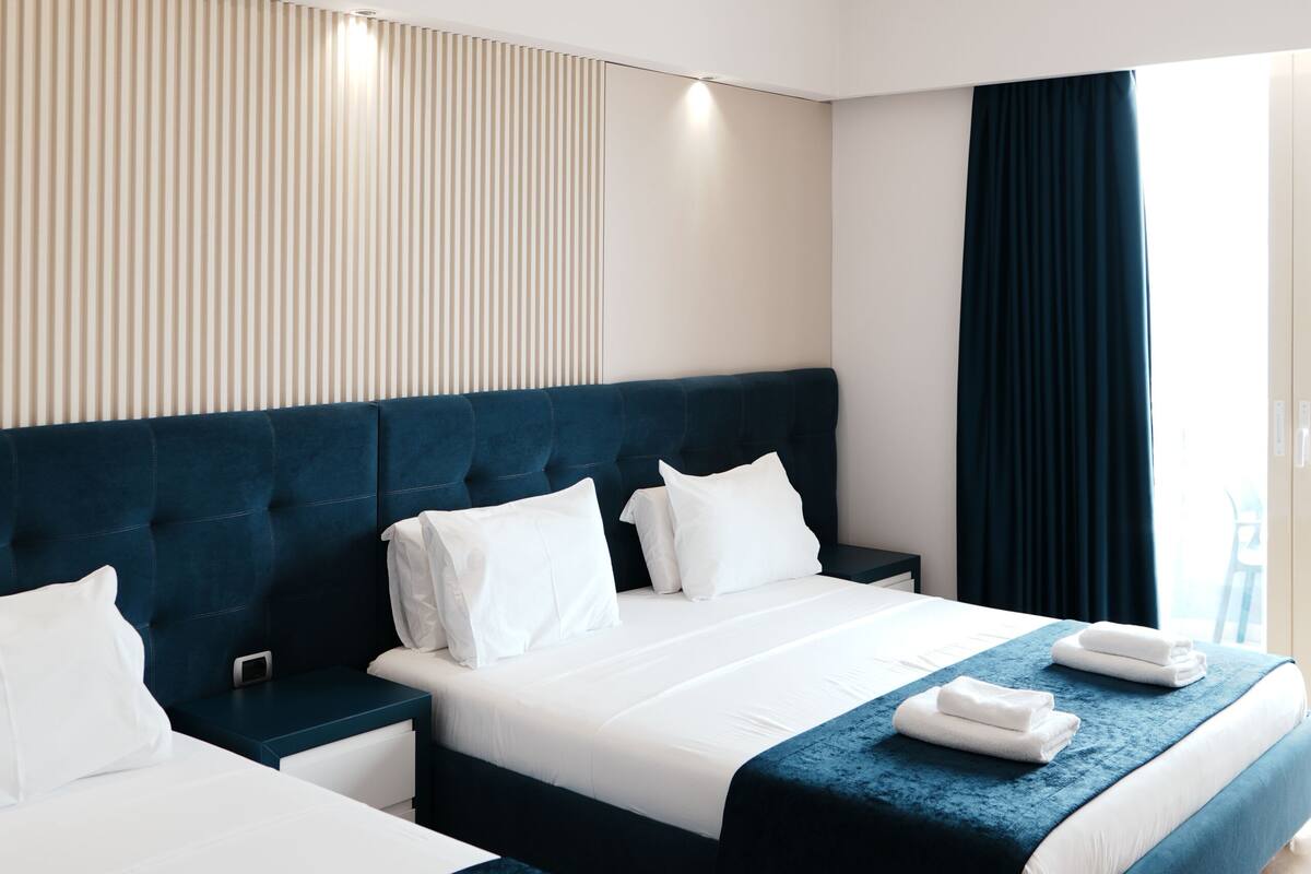 A hotel room with two beds, a large window with curtains, a ceiling light, and a wall-mounted panel.