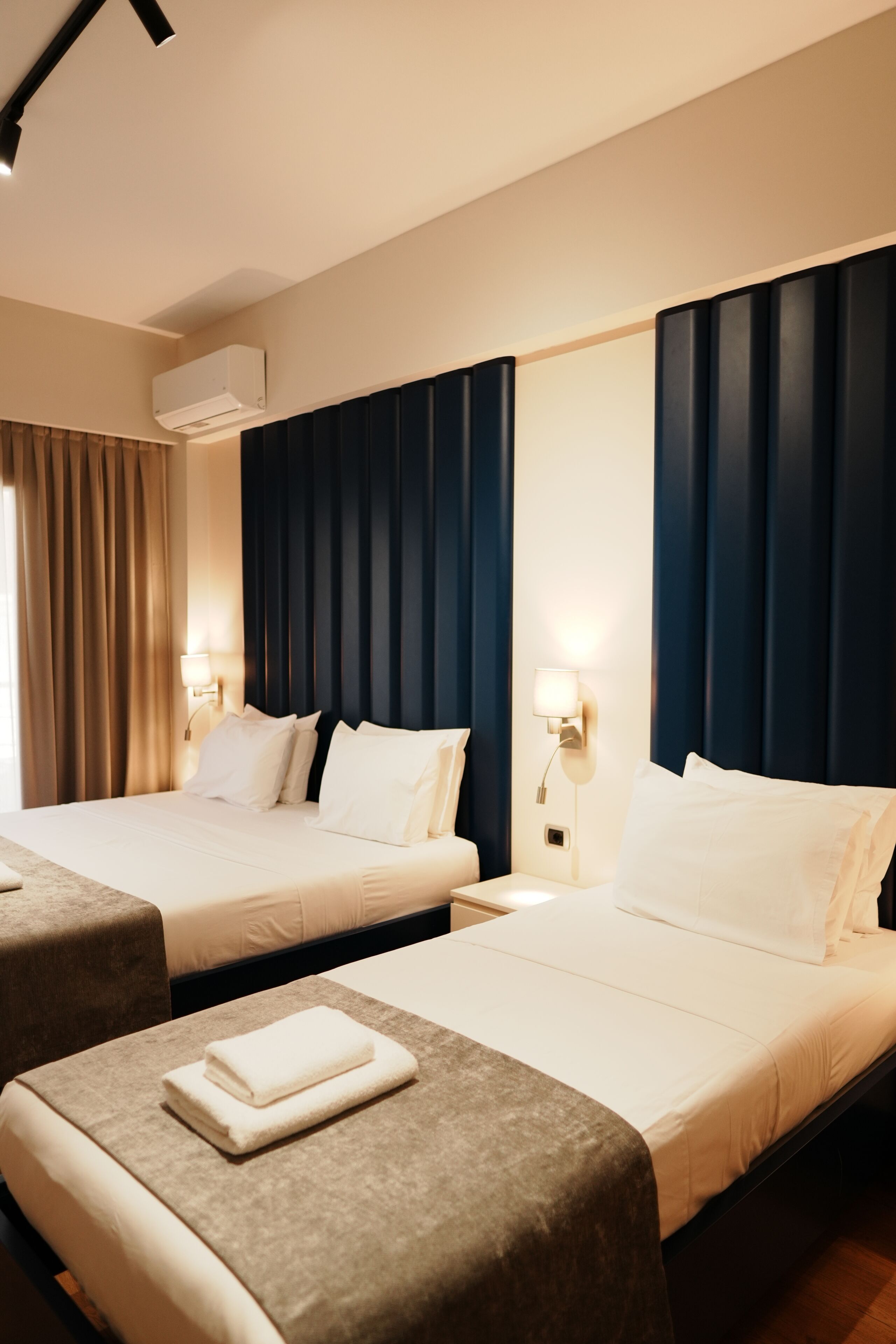 A hotel room with two beds, a wall-mounted air conditioner, bedside lamps, and a window with curtains.