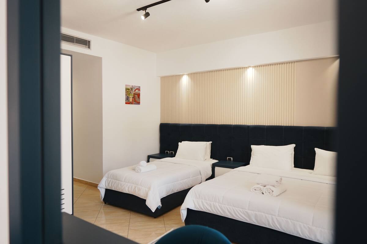A hotel room with two beds, a large window with blinds, a small artwork on the wall, and a door leading to another room.