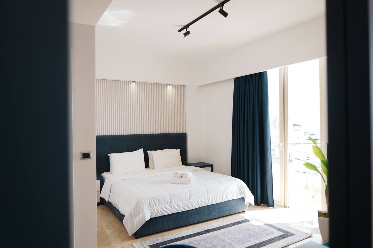 A modern bedroom with a large bed, a dark headboard, and a view of the outdoors through a large window.