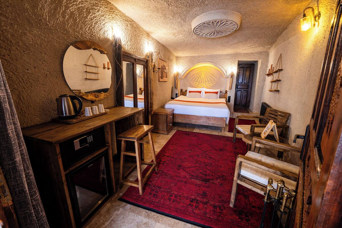 A cozy room with a bed, a desk with a kettle, a chair, and a red rug.