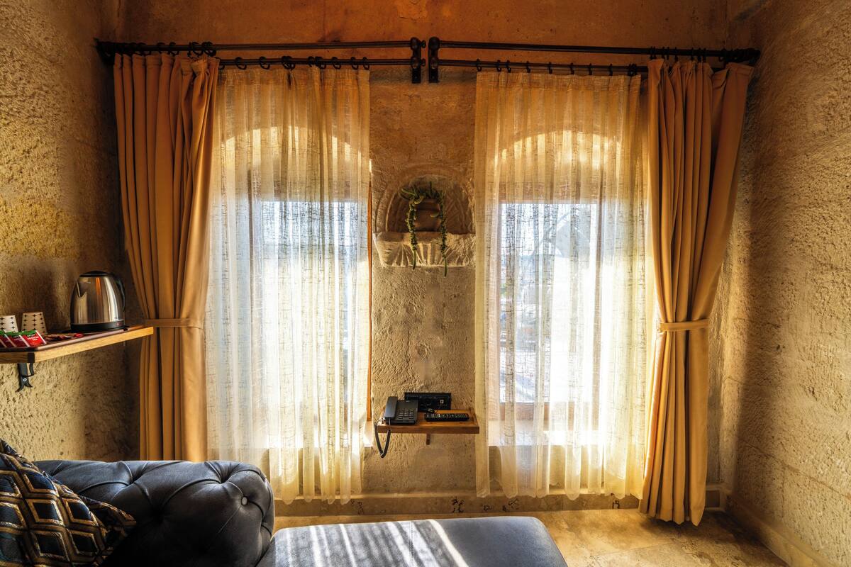 A room with a bed, a small shelf with a kettle and a phone, and a window with curtains.