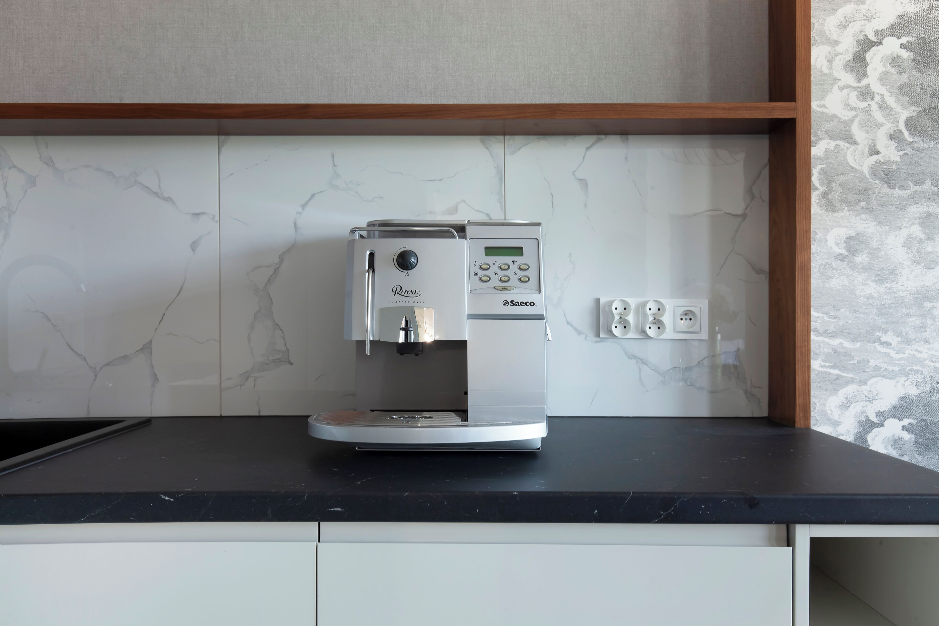 Cozinha moderna com cafeteira embutida em bancada preta, armários brancos e painel de backsplash em mármore.
