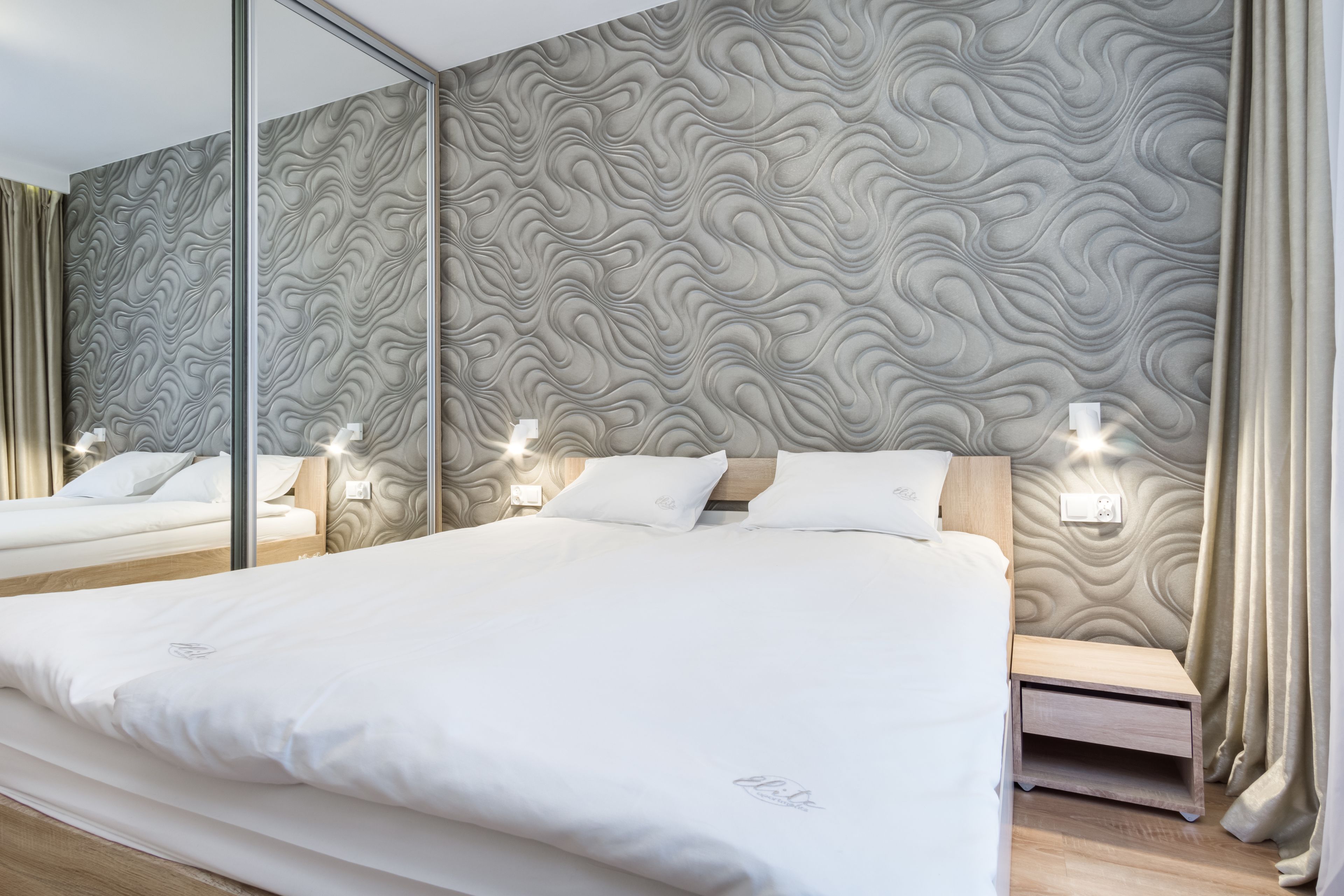 Une chambre moderne avec un grand lit, une table de chevet, un miroir et un mur orné d’un motif ondulé.