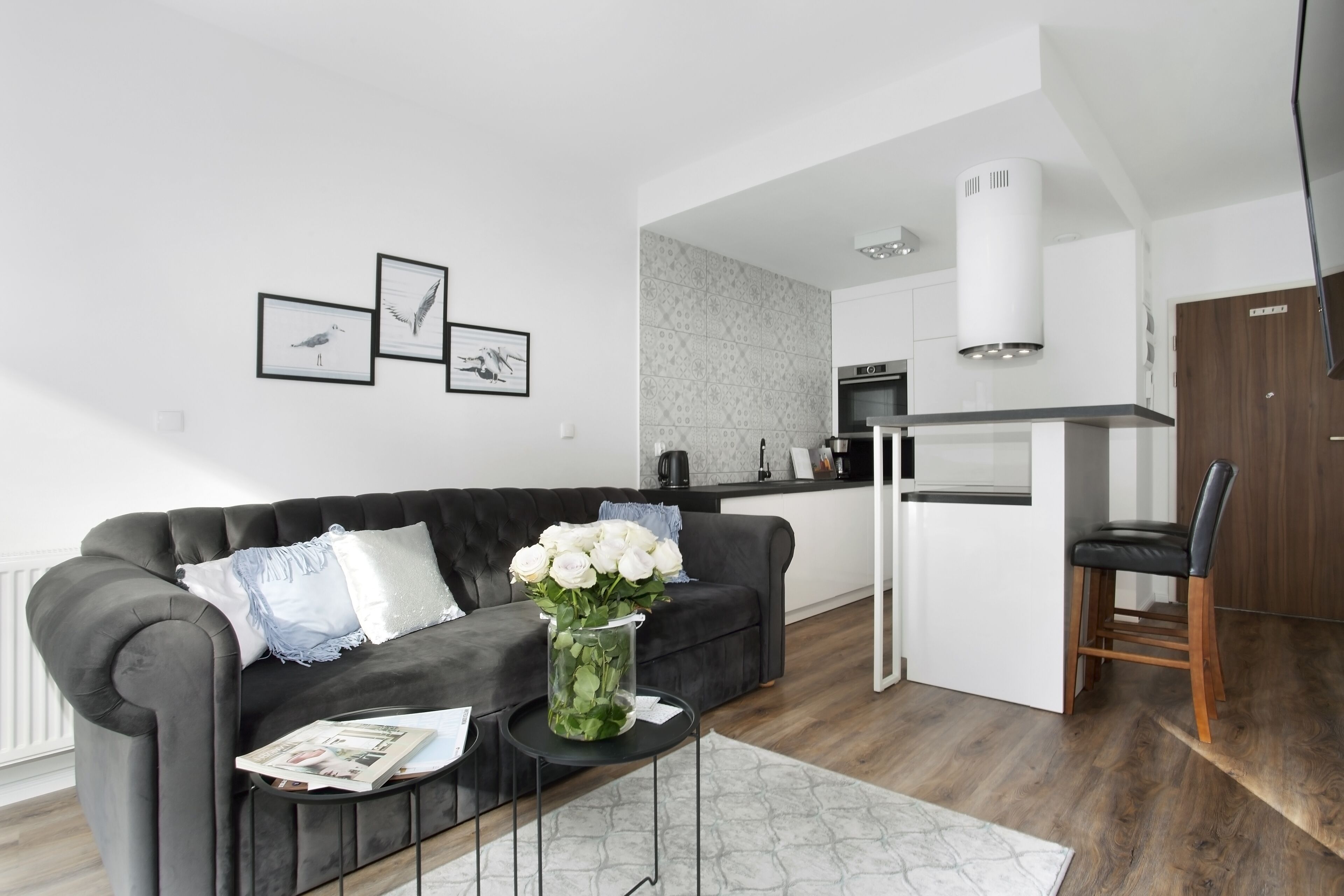 Un salon moderne avec un canapé gris foncé, une petite table ronde ornée d’un vase de fleurs blanches, et une cuisine en arrière-plan.