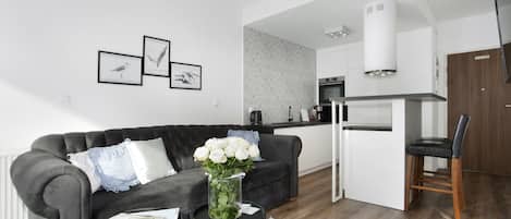 Un salon moderne avec un canapé gris foncé, une petite table ronde ornée d’un vase de fleurs blanches, et une cuisine en arrière-plan.