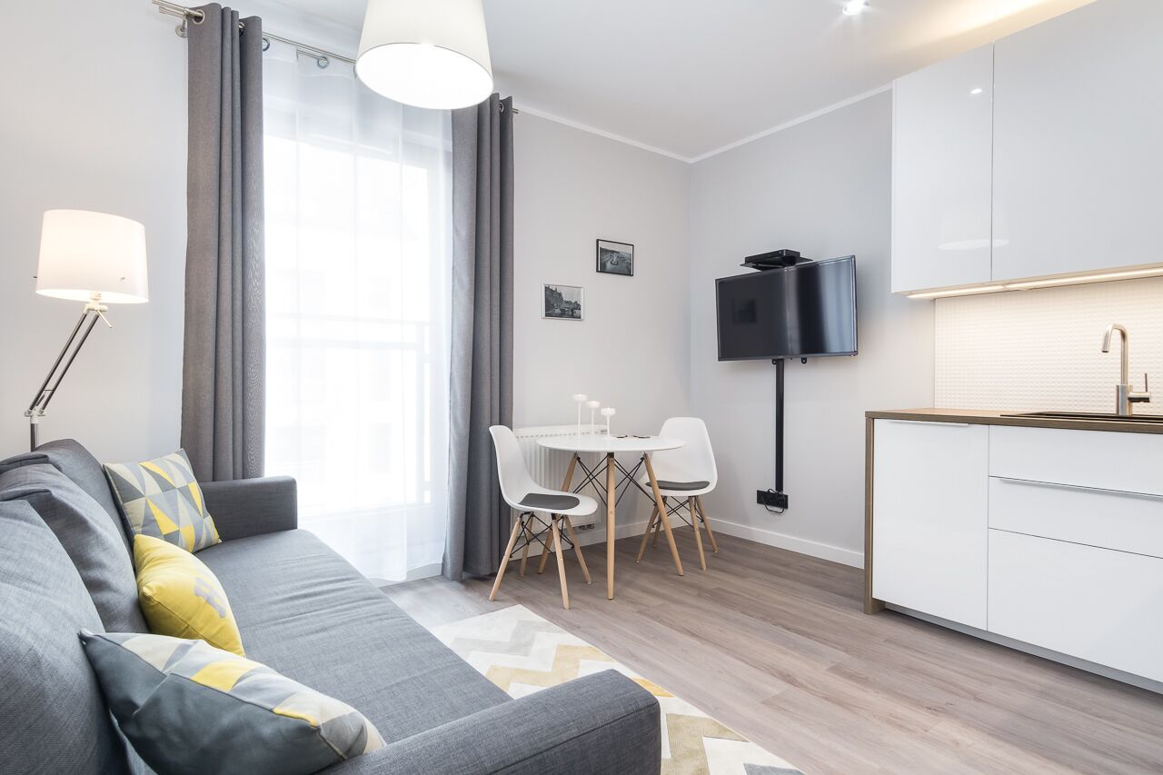 A modern living room with a grey sofa, a dining table with chairs, a flat-screen TV mounted on the wall, and a kitchen area with white cabinets and a sink.