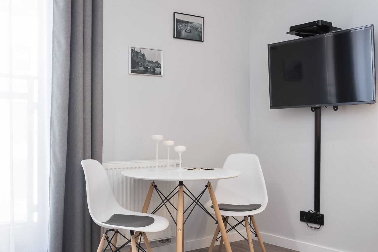 Een moderne kamer met een ronde witte tafel, twee witte stoelen, een flatscreen televisie aan de muur en twee ingekaderde foto's.