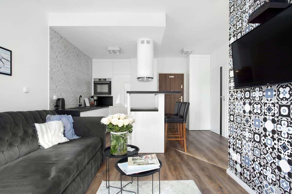 Un salón moderno con un sofá gris oscuro, una mesita de centro con una revista y un jarrón con flores blancas. Se ve una cocina con armarios blancos y encimera negra al fondo.