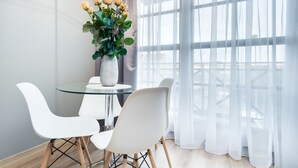 A modern dining area with a glass table and white chairs, a vase with yellow flowers, and a view through sheer curtains. - Elite Apartments Spa Zone (Gdansk)