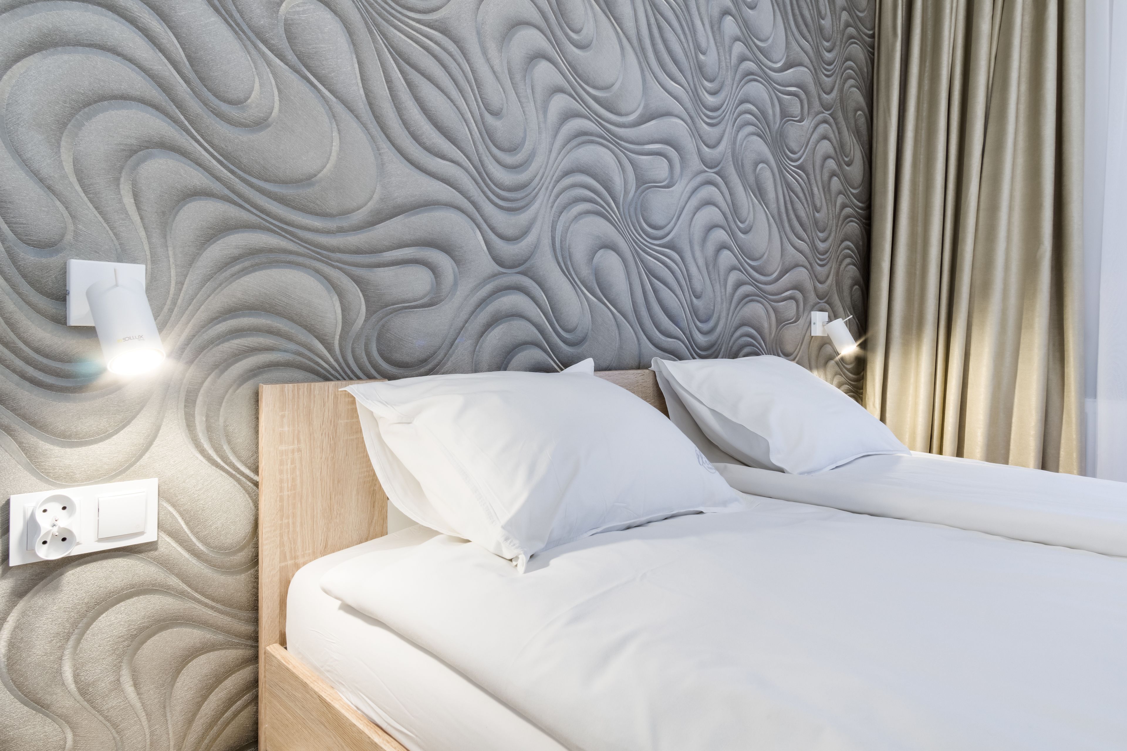 A neatly made bed with white linens and pillows, a wall with a textured pattern, a bedside lamp, and a power outlet.