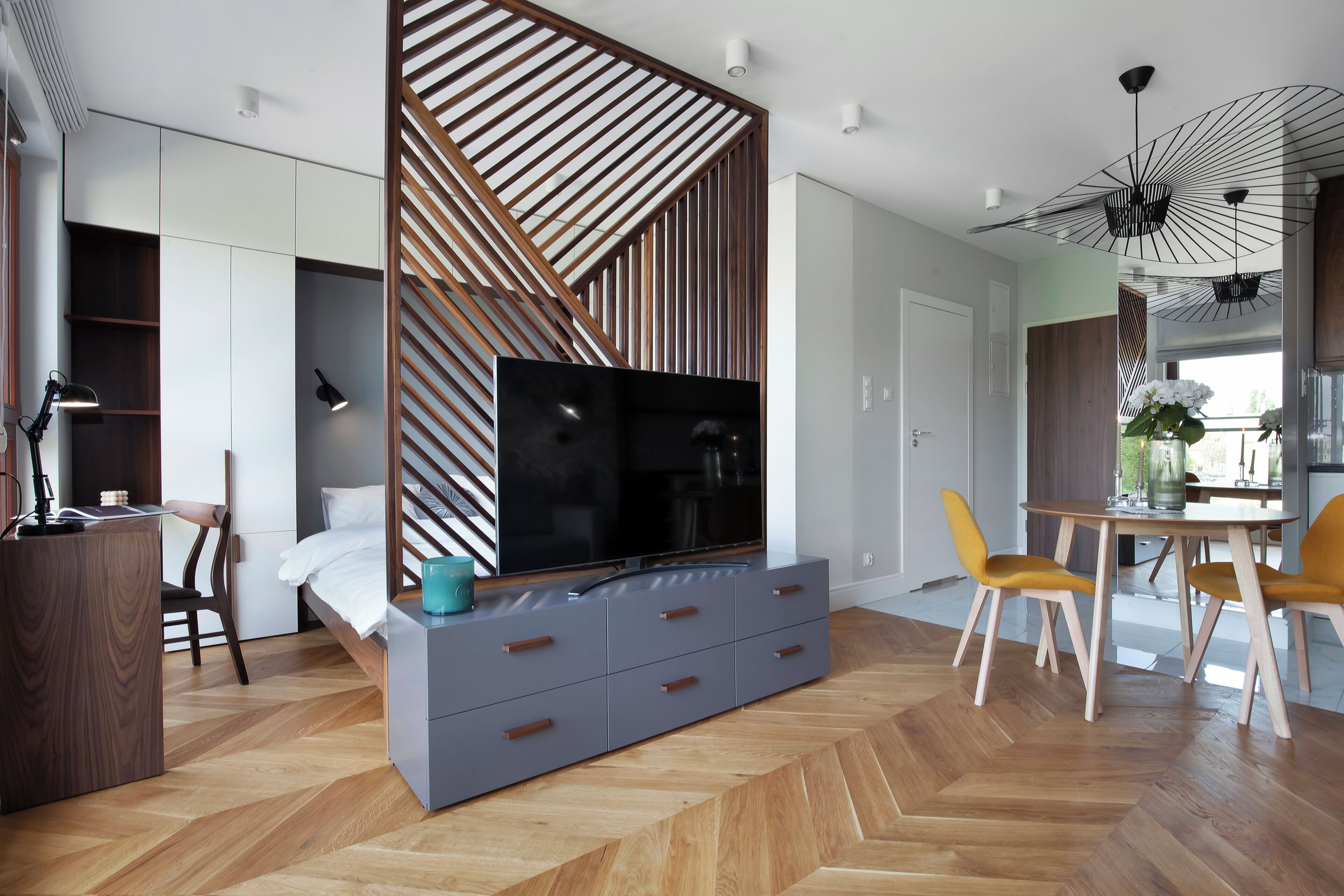 Un salon moderne doté d’un téléviseur, d’une cloison en lattes de bois, de meubles de rangement gris, d’un parquet en bois, de chaises jaunes et d’un coin repas avec une table et des chaises.