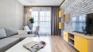 A modern living room with a grey sofa, a white coffee table with magazines, a dining area with a glass table and chairs, and a TV on a wooden cabinet. - Elite Apartments Spa Zone (Gdansk)