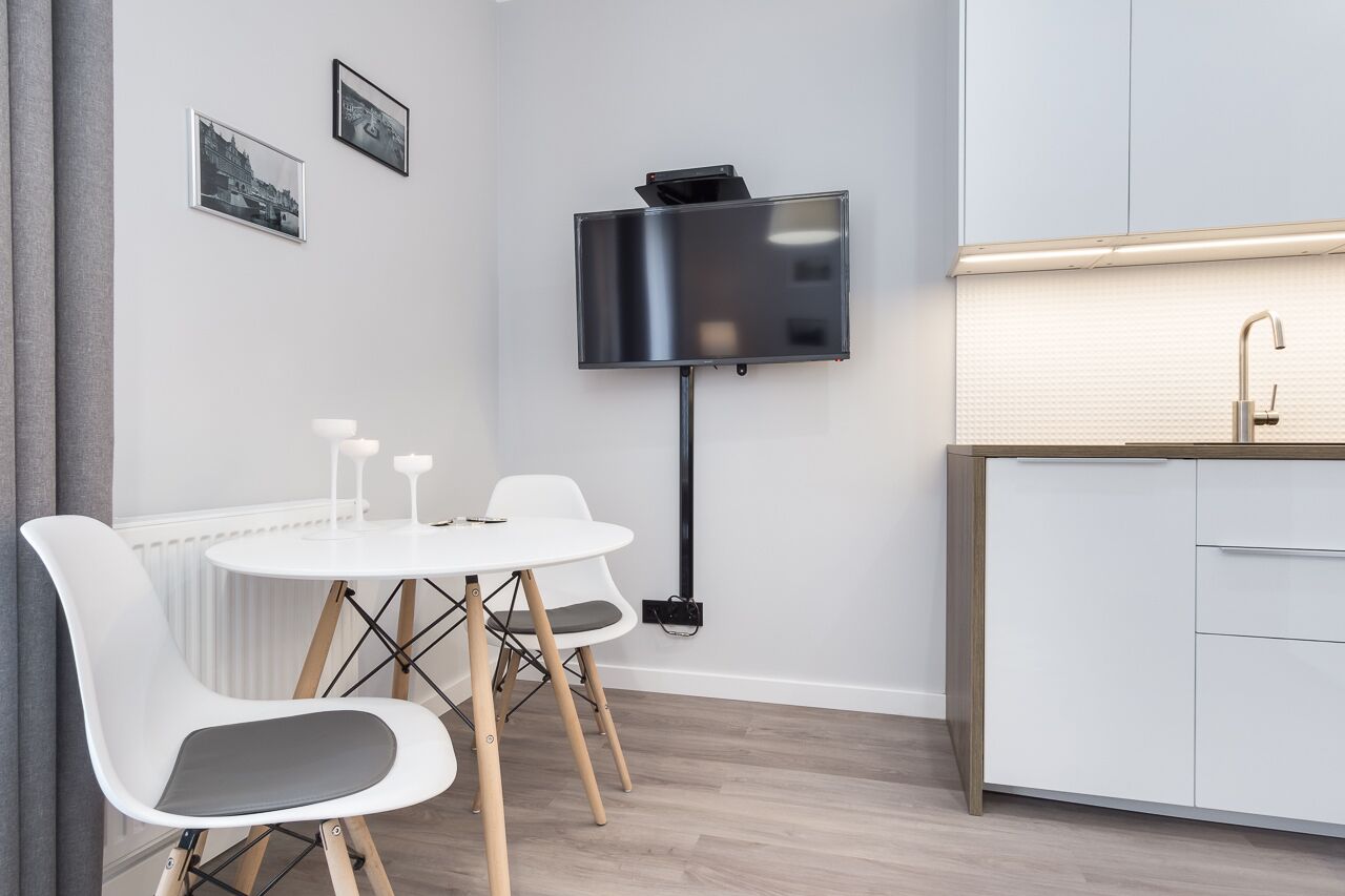 A modern kitchen with a built-in sink, a flat-screen TV mounted on the wall, and a dining area with a round table and white chairs.
