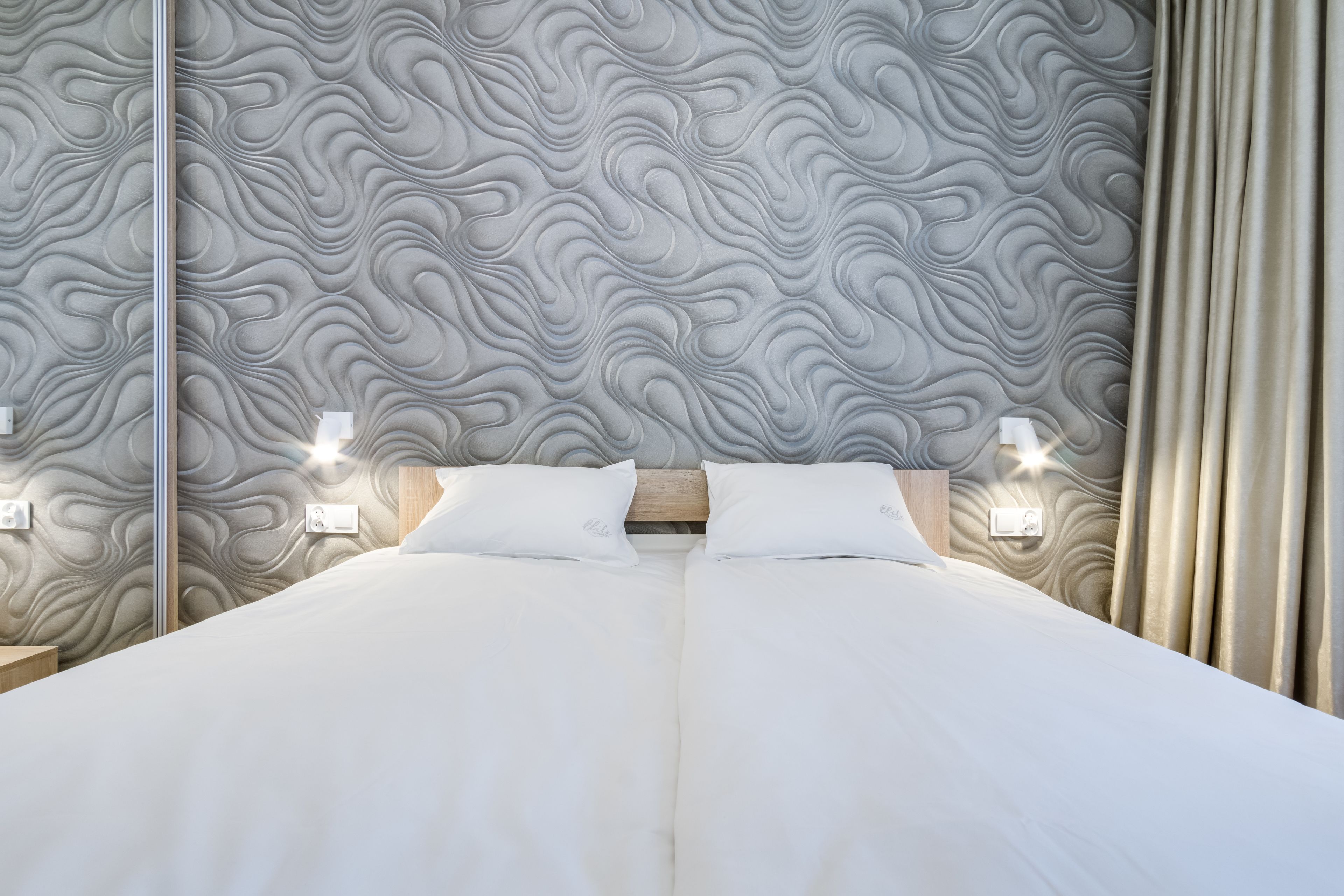 Un dormitorio moderno con una cama grande, cabecera con diseño ondulado, lámparas de noche y un espejo en la pared.