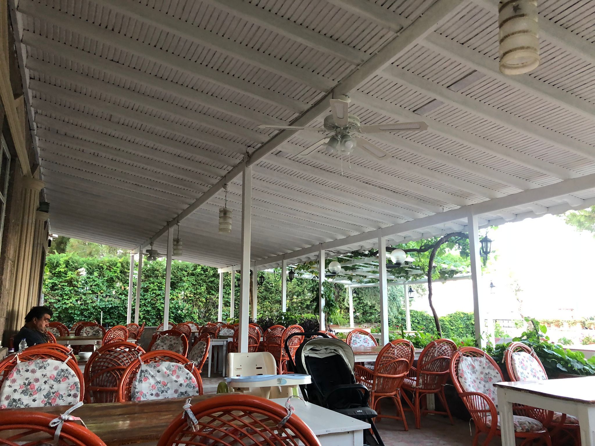 An outdoor cafe with wicker chairs and tables, a white ceiling, and greenery in the background.