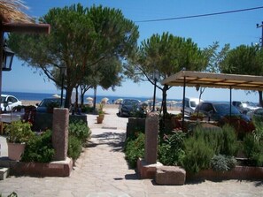 Un chemin pavé mène à une zone située au bord de la plage, où sont garées des voitures et où se trouvent des tables et des chaises à l’extérieur, sous des parasols.