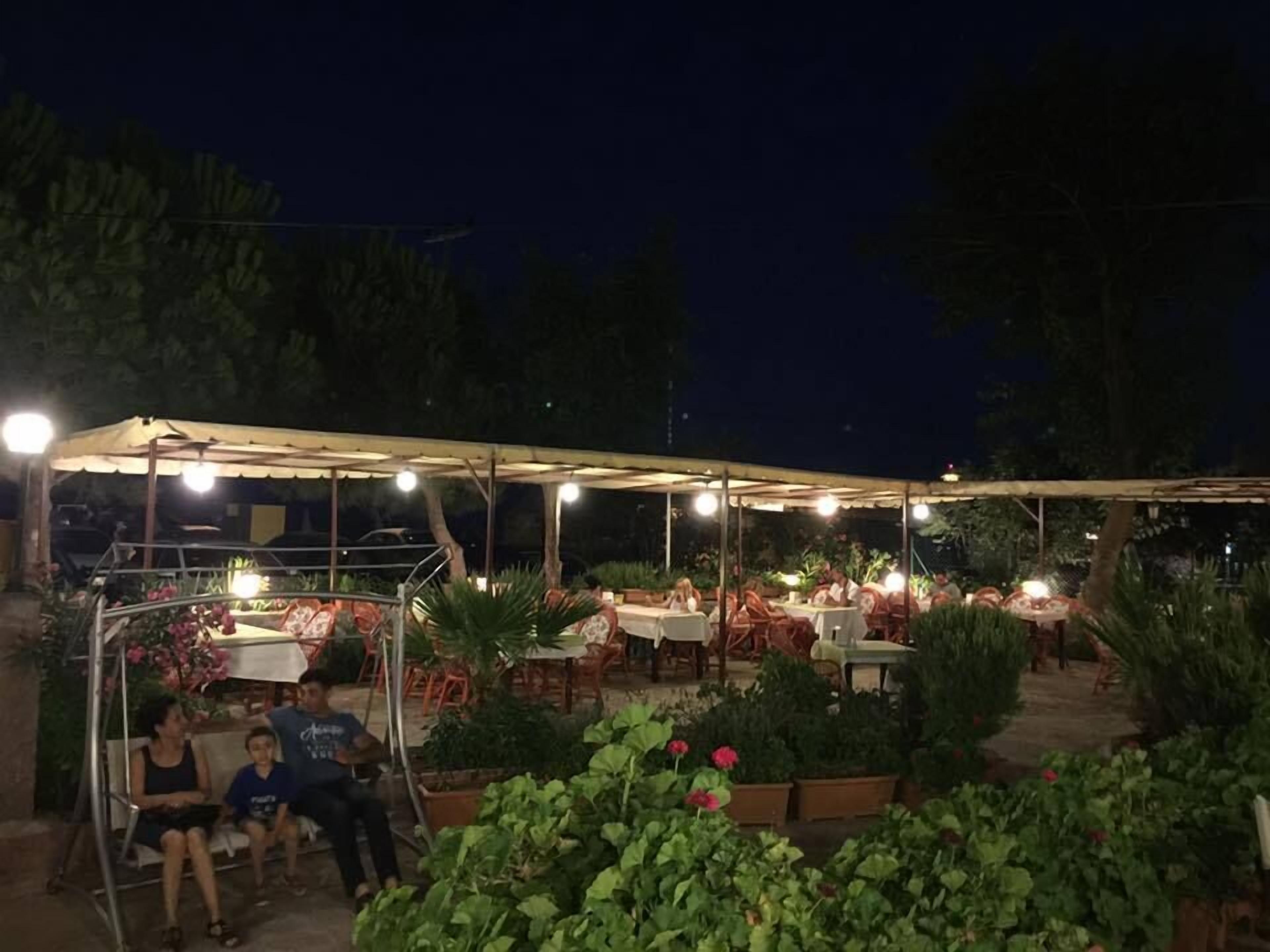An outdoor dining area with tables and chairs under a covered structure, surrounded by potted plants and greenery.