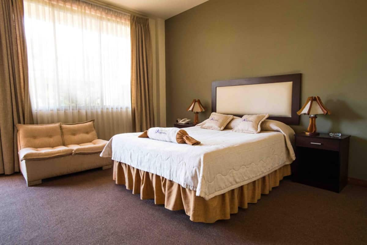 A hotel room with a bed, a sofa, two bedside lamps, a nightstand, and a window with curtains.