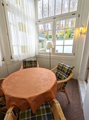 Una habitación acogedora con una mesa redonda cubierta por un mantel color melocotón, flanqueada por sillas de mimbre con cojines a cuadros. Una lámpara de pie se encuentra cerca de la ventana, que ofrece vistas de una zona residencial.