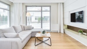 Una sala de estar moderna con un sofá de color claro, una mesa de centro de madera y una lámpara de pie. Hay un ventanal con persianas, un televisor de pantalla plana montado en la pared y una pequeña planta en maceta sobre una estantería.