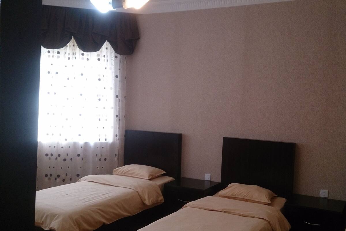 A hotel room with two single beds, a small table, and a window with curtains.
