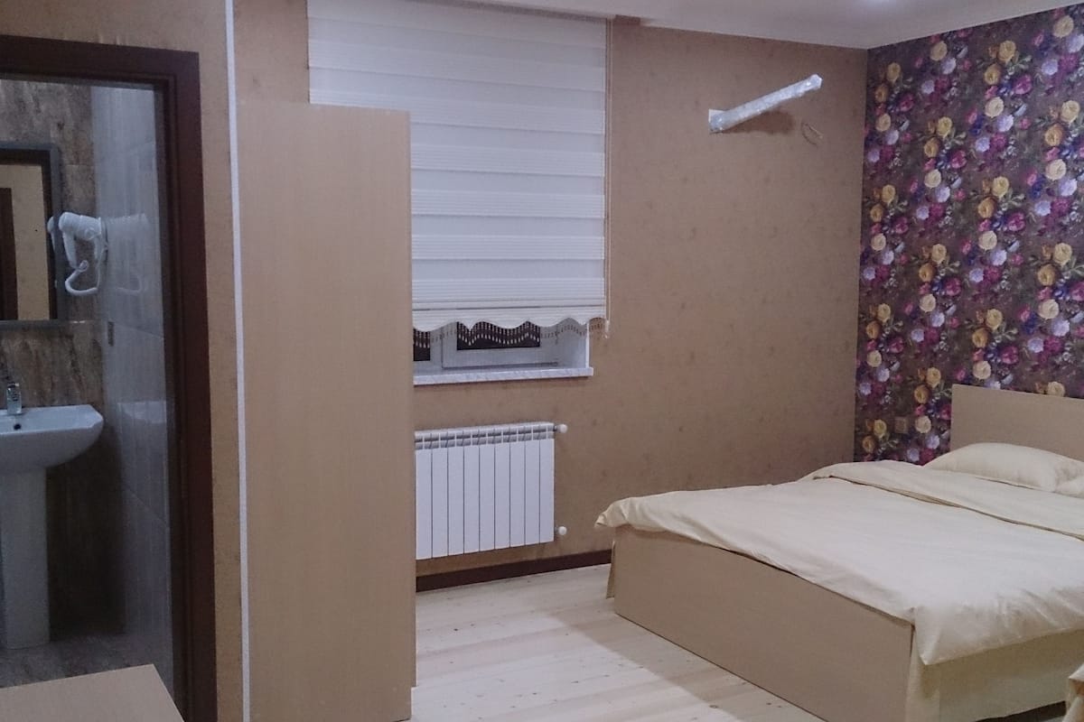 A bedroom with a bed, a window with blinds, a radiator, and a bathroom with a sink and mirror.