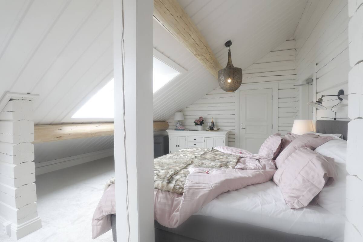 A bedroom with a sloped ceiling, a bed with pink bedding, a wooden beam, and a hanging light fixture.