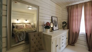 A bedroom with a large mirror, a tufted chair, a vanity with drawers, a bed with pillows, and a window with curtains. - Sara's Bed and Breakfast (Erikslund)