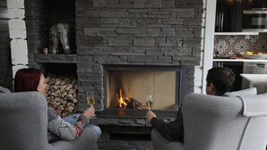 A cozy living room with a stone fireplace, two people seated facing the fire, and a small table with books and a glass. - Sara's Bed and Breakfast (Erikslund)