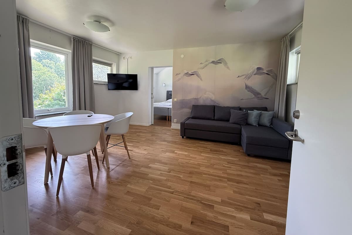Una sala de estar moderna con piso de madera, un sofá, una mesa de comedor con sillas, un televisor montado en la pared y un mural de pájaros.