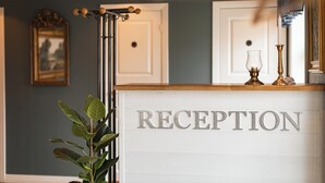 A reception desk with 'RECEPTION' signage, a coat rack, a framed picture on the wall, and a potted plant. - Hensbacka Hotell (Munkedal)