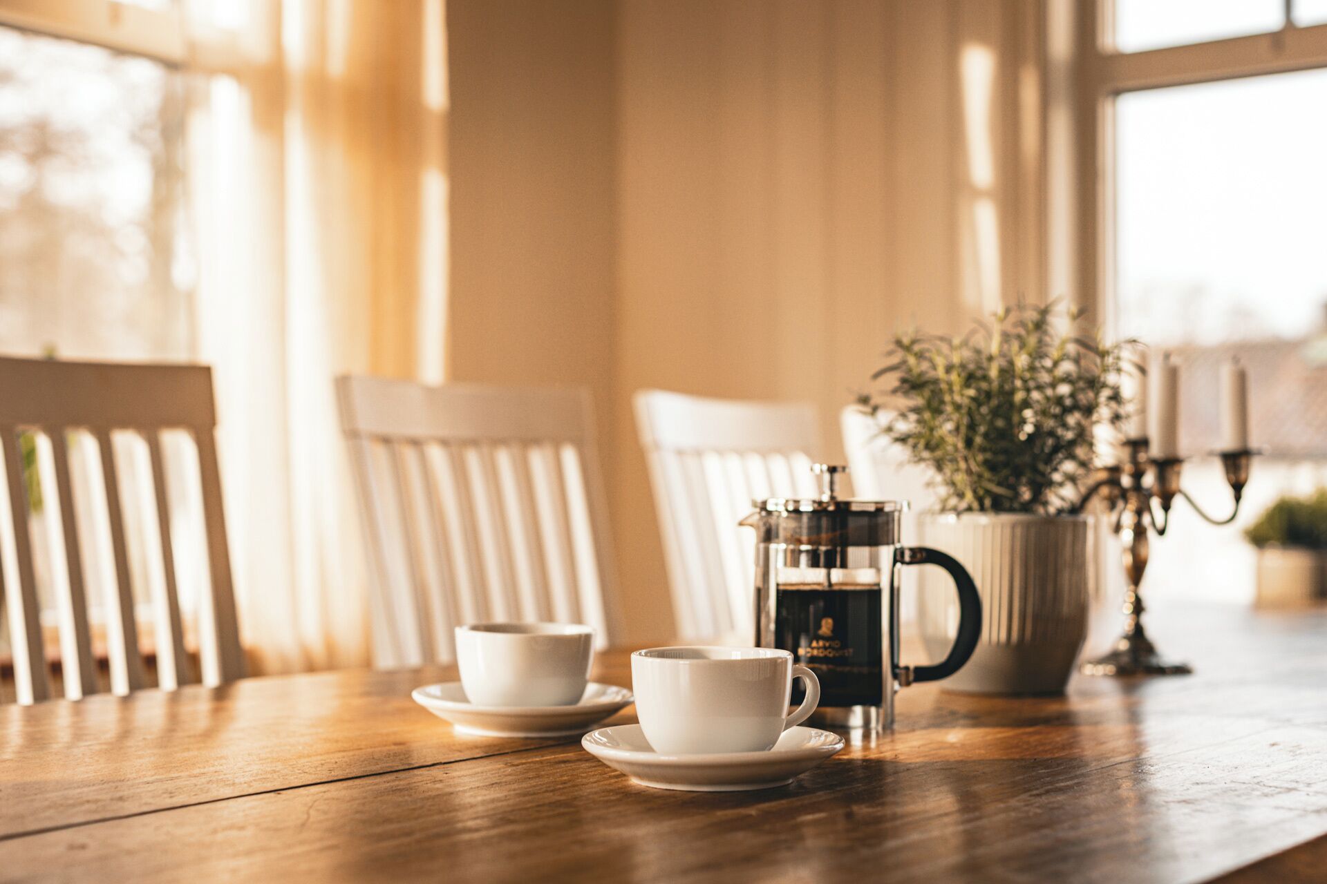 Uma mesa de madeira com duas xícaras brancas e uma cafeteira, uma planta em vaso e um candelabro.