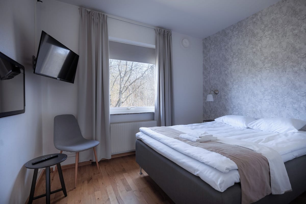 Un dormitorio con cama, una silla, un televisor montado en la pared y una ventana con cortinas.
