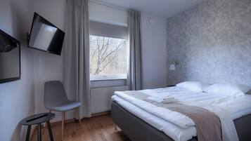 Un dormitorio con cama, una silla, un televisor montado en la pared y una ventana con cortinas.