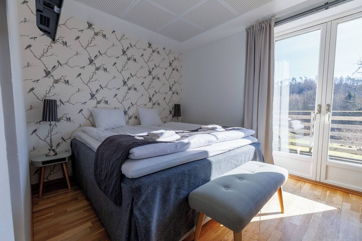 Un dormitorio con cama, banqueta, mesita de noche, lámpara y ventana con vistas.