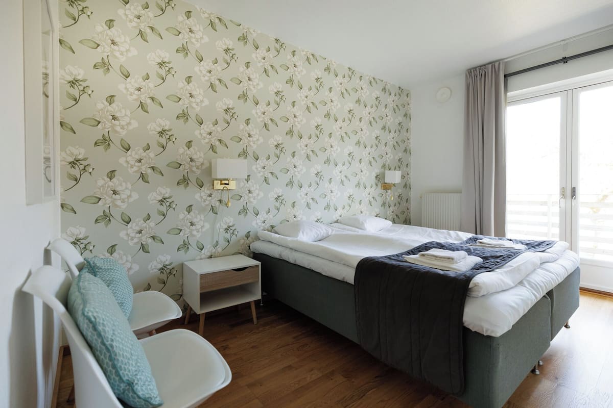 Un dormitorio con papel tapiz floral, una cama con sábanas blancas, una mesita de noche y una lámpara de noche.