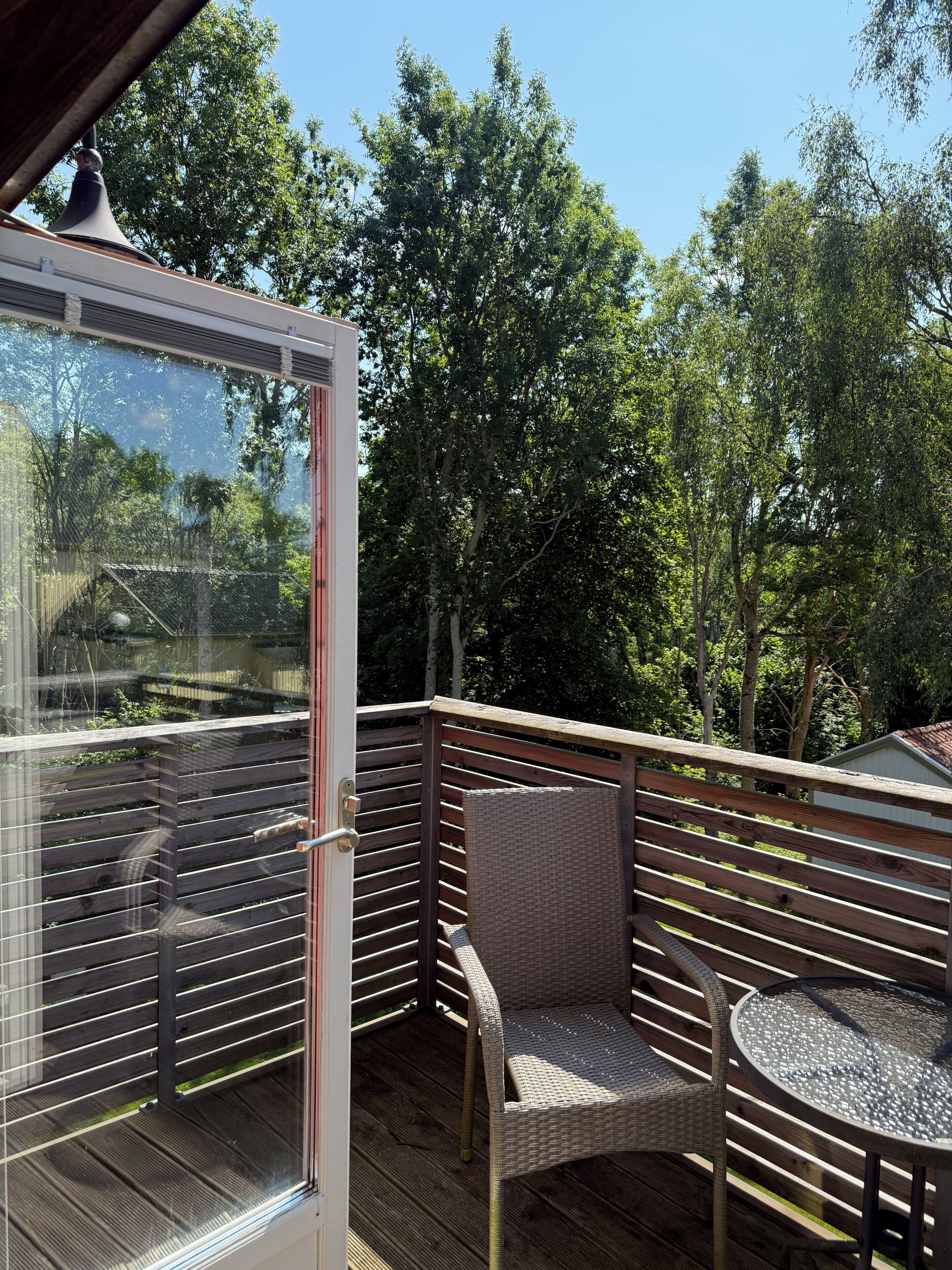 A covered outdoor seating area with a glass door, wooden deck, wicker chairs, and a small round table.