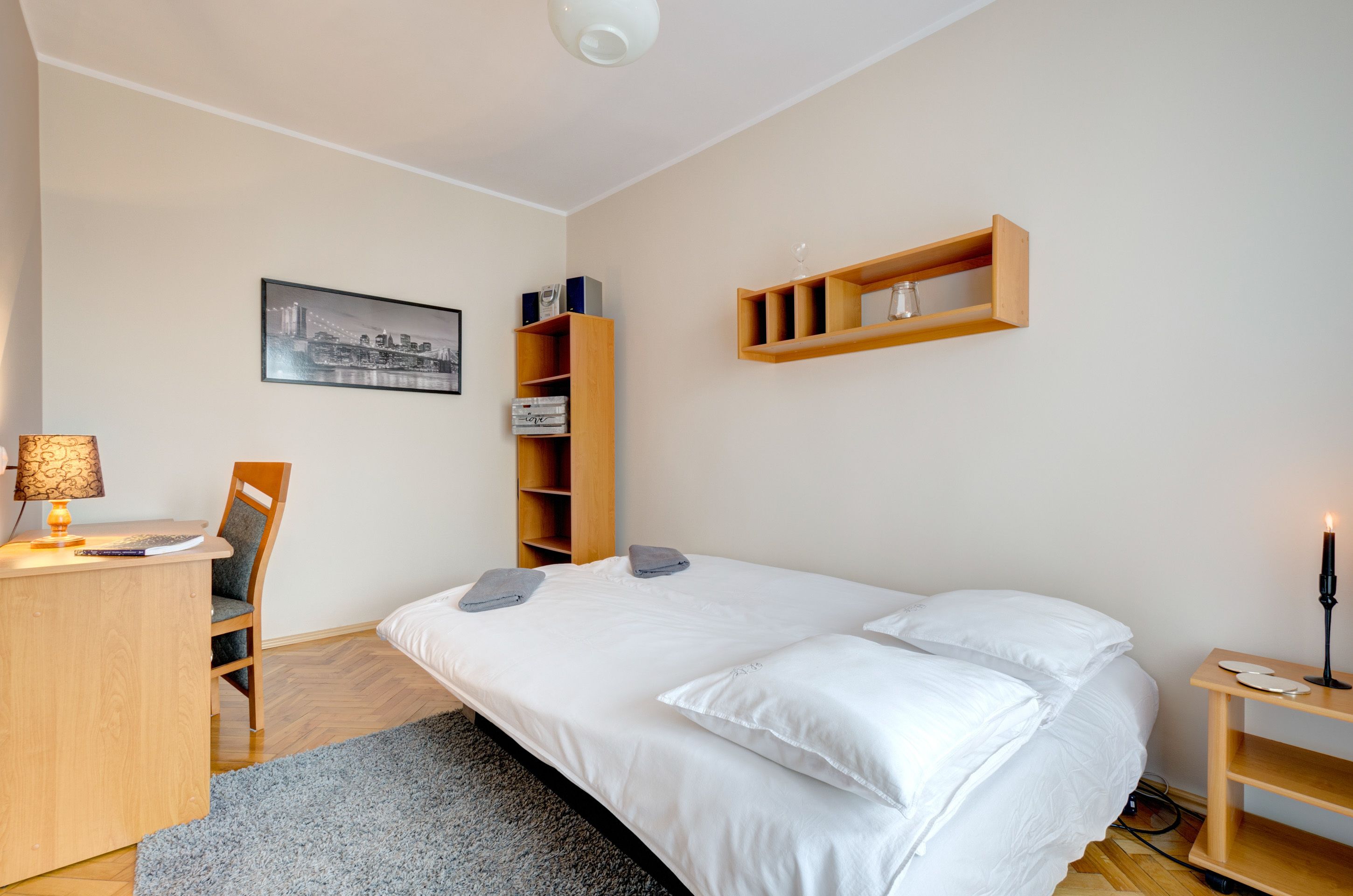 A neatly arranged bedroom with a bed, a desk, a chair, a bookshelf, and a bedside table with a lamp.