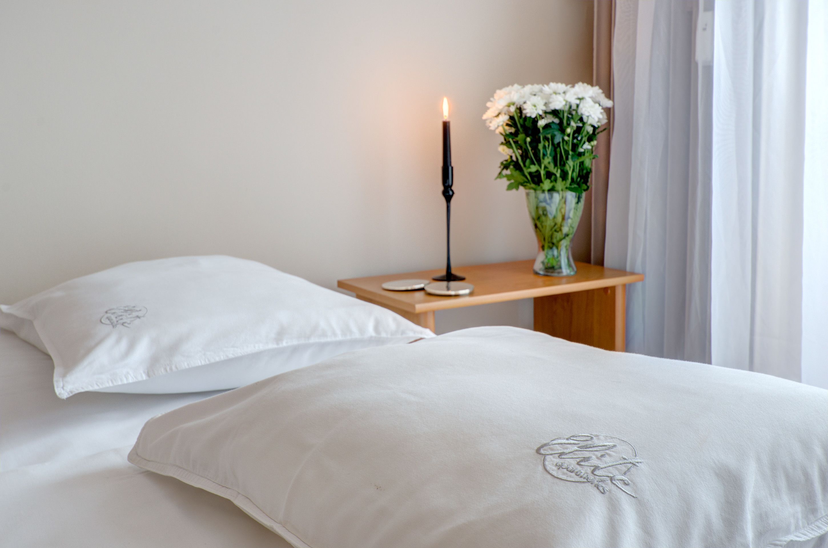 A bedroom with two white beds, a bedside table with a candle, and a vase with white flowers.