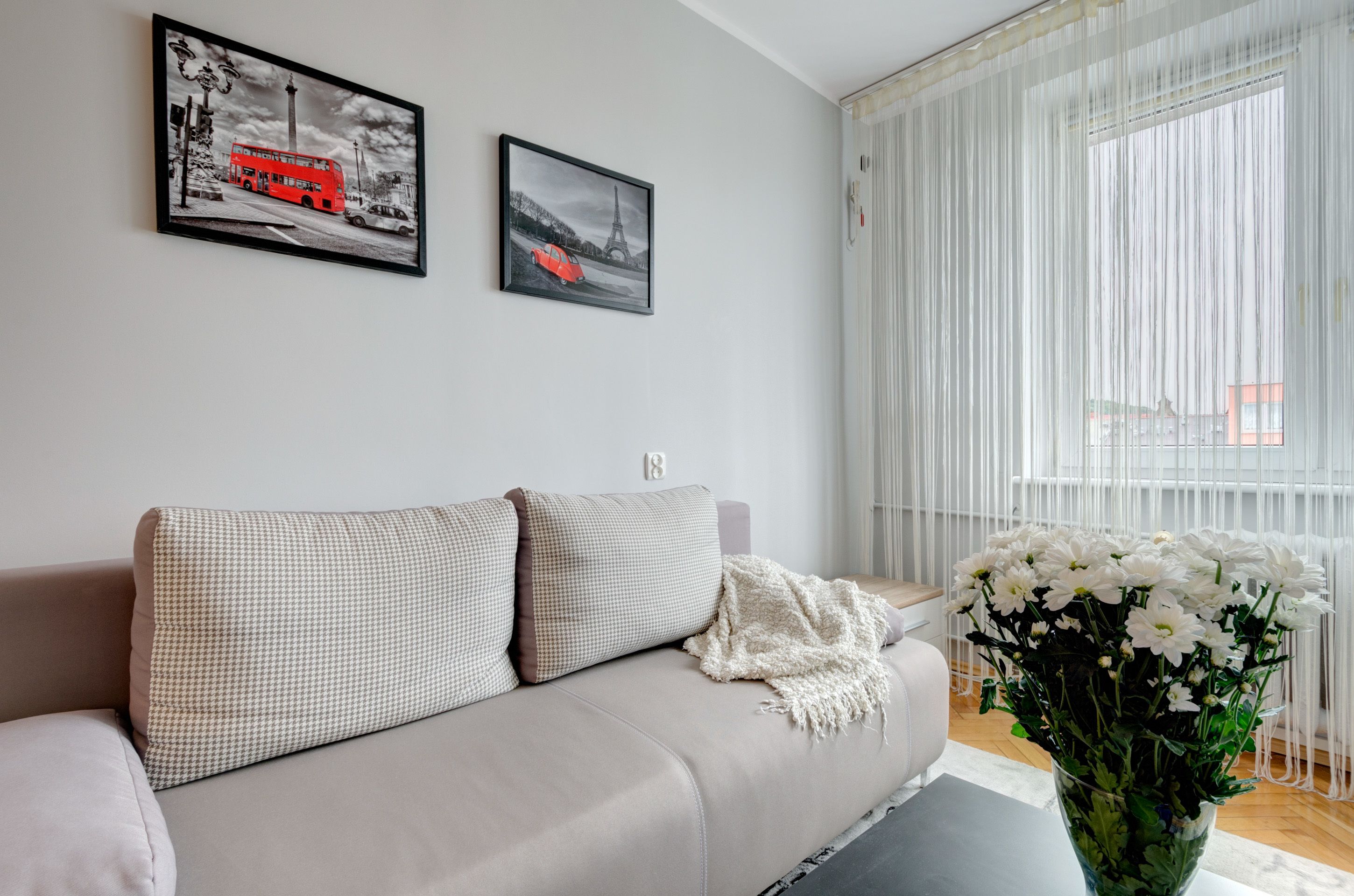 A living room with a beige sofa, two framed pictures on the wall, and a vase of white flowers.