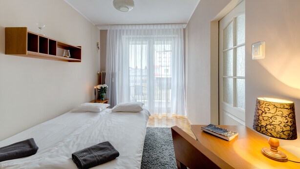 Quarto com duas camas, uma escrivaninha, luminária e janela com cortinas leves.