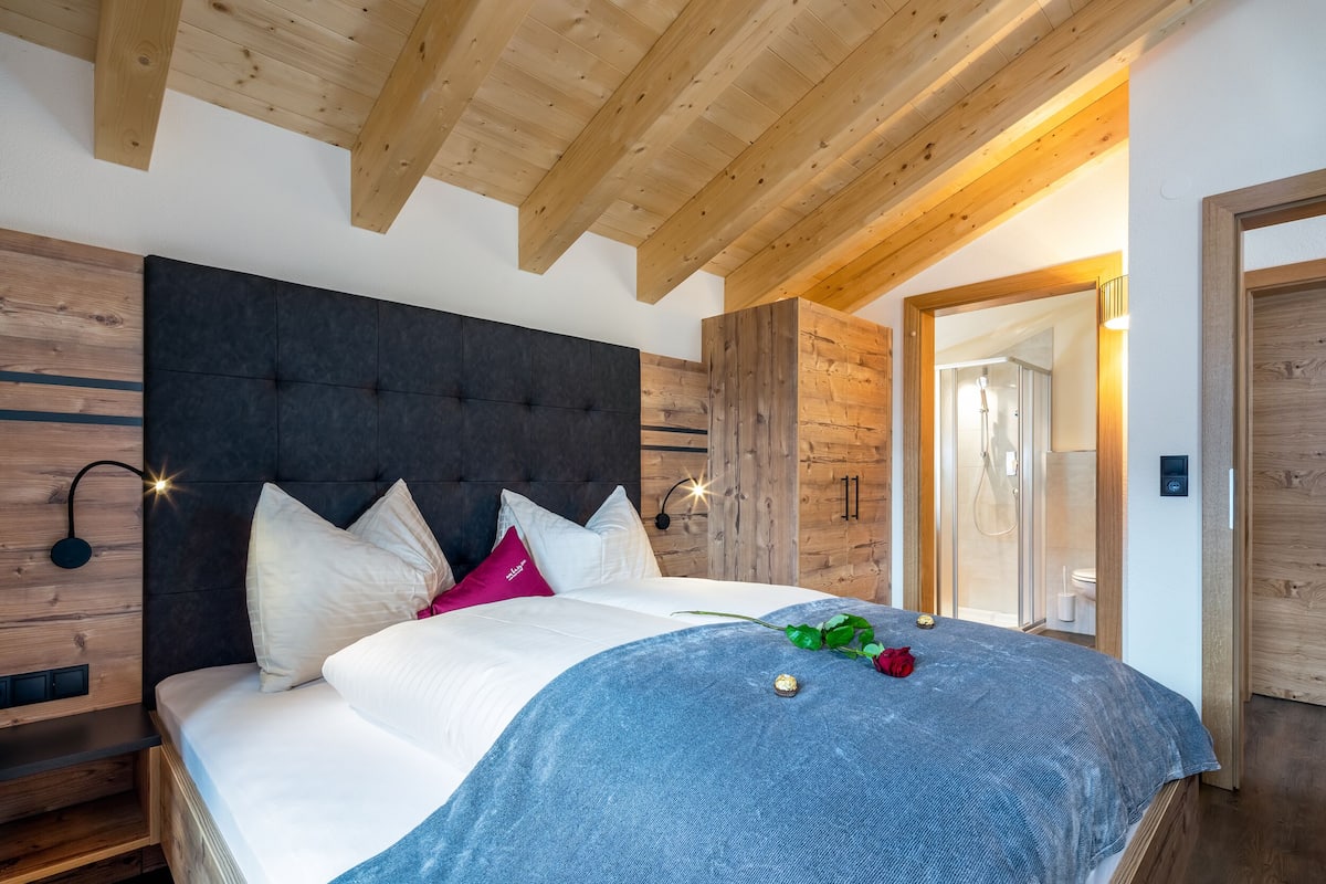 A bedroom with a large bed, wooden ceiling beams, a dark headboard, and a wooden wardrobe.