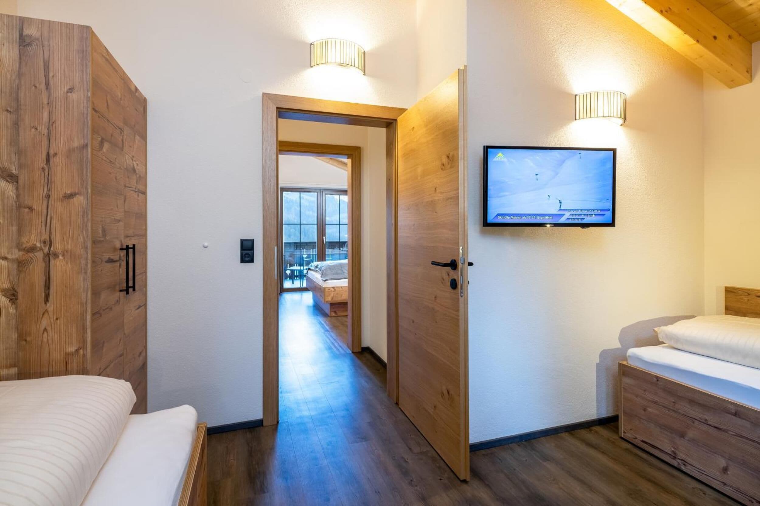 A modern hotel room with a wooden wardrobe, a bed, a TV mounted on the wall, and a hallway leading to another room.