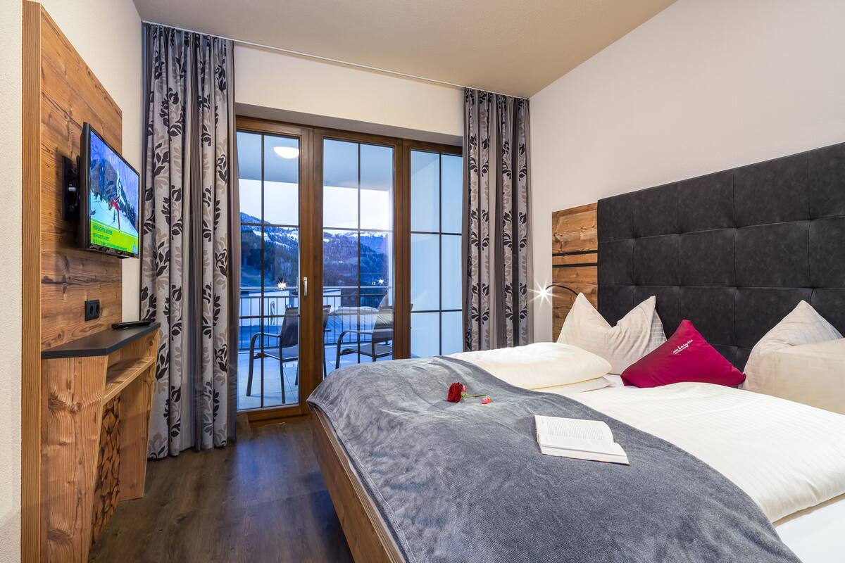 A hotel room with a large bed, a TV on the wall, a wooden desk, and a view of the outdoors through the window.