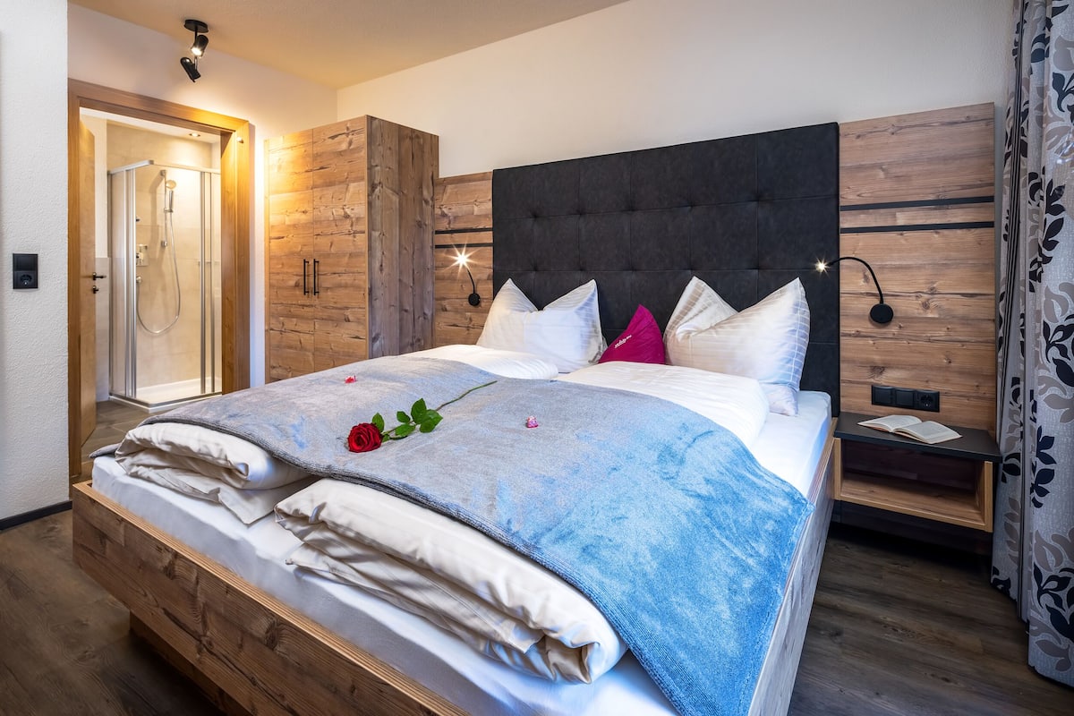 A bedroom with a wooden headboard, a bed with white and blue bedding, a rose decoration, and a nightstand with an open book.