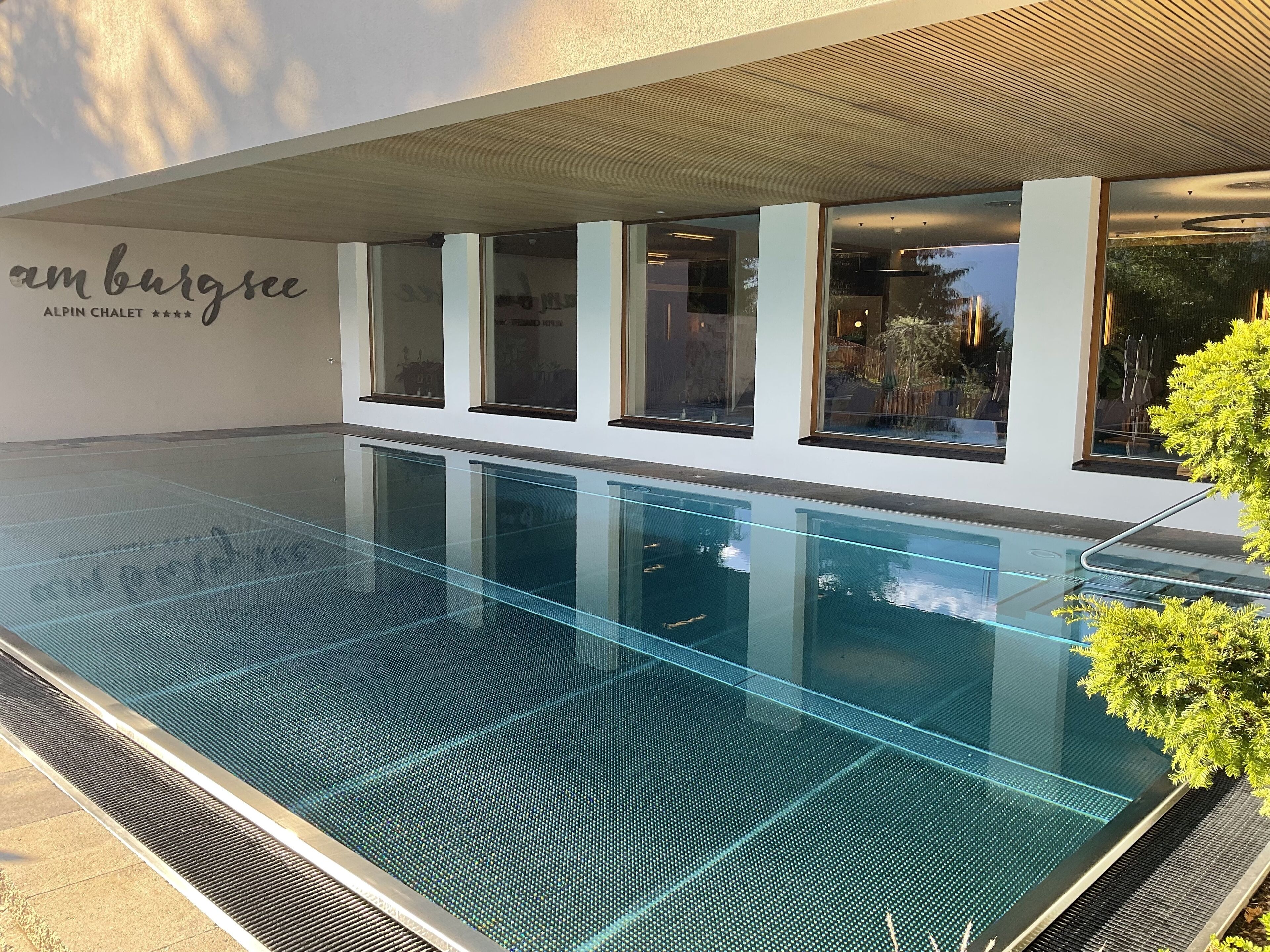 Un bâtiment moderne doté d’une piscine à fond de verre, de grandes fenêtres et d’une enseigne indiquant « Amburgsee ALPIN CHALET ».
