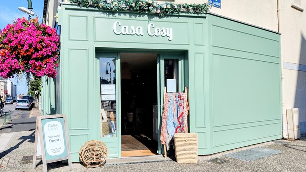 A storefront named "Casa Cosy" with a green facade, a hanging flower basket, and a wicker chair outside.