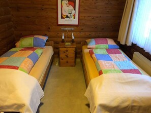 A cozy cabin-style room with two beds, each with colorful quilts, a wooden nightstand with a lamp, and a framed artwork on the wall.